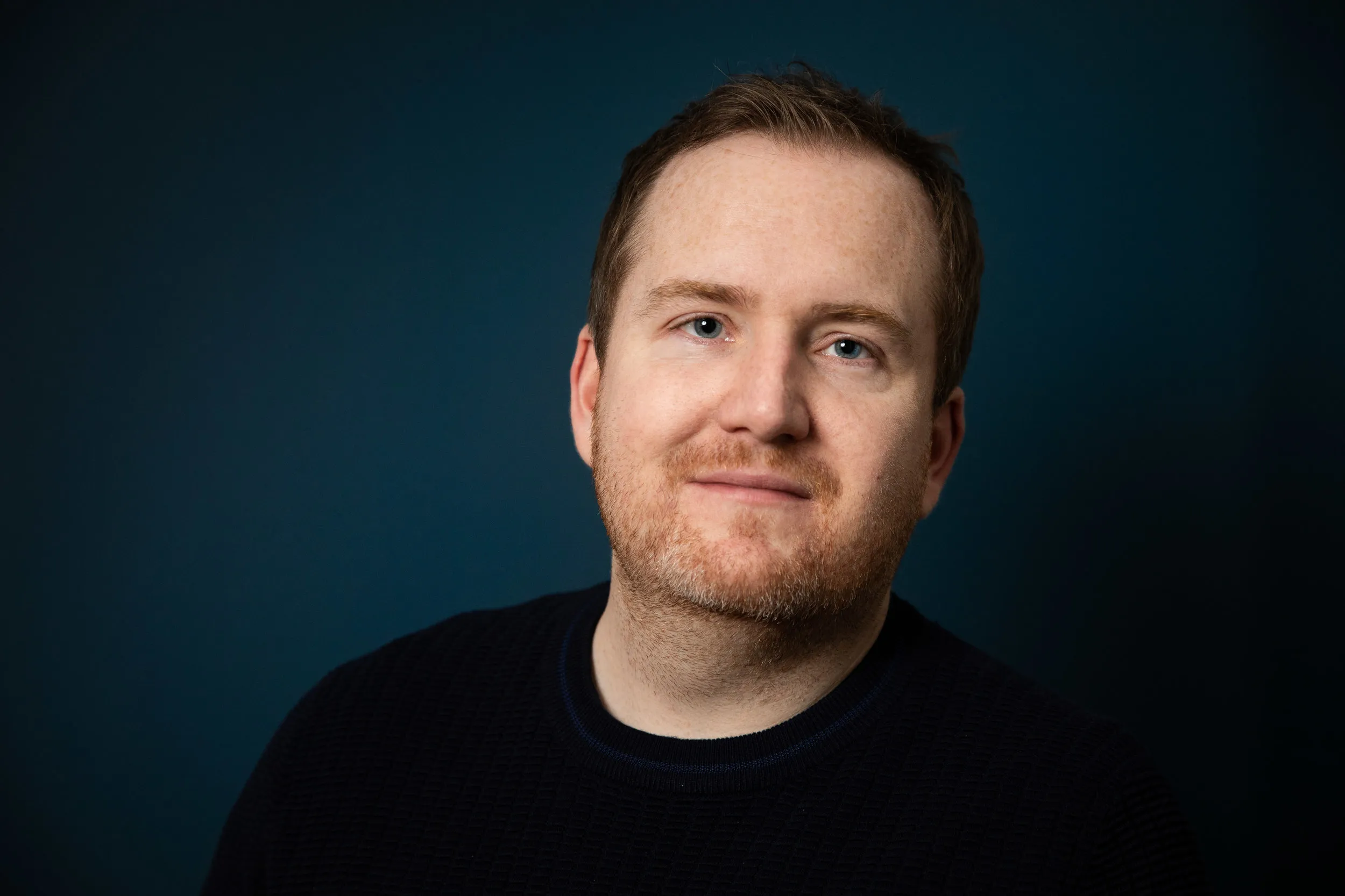 Corporate headshot of man with ginger beard in navy sweater, teal background, London