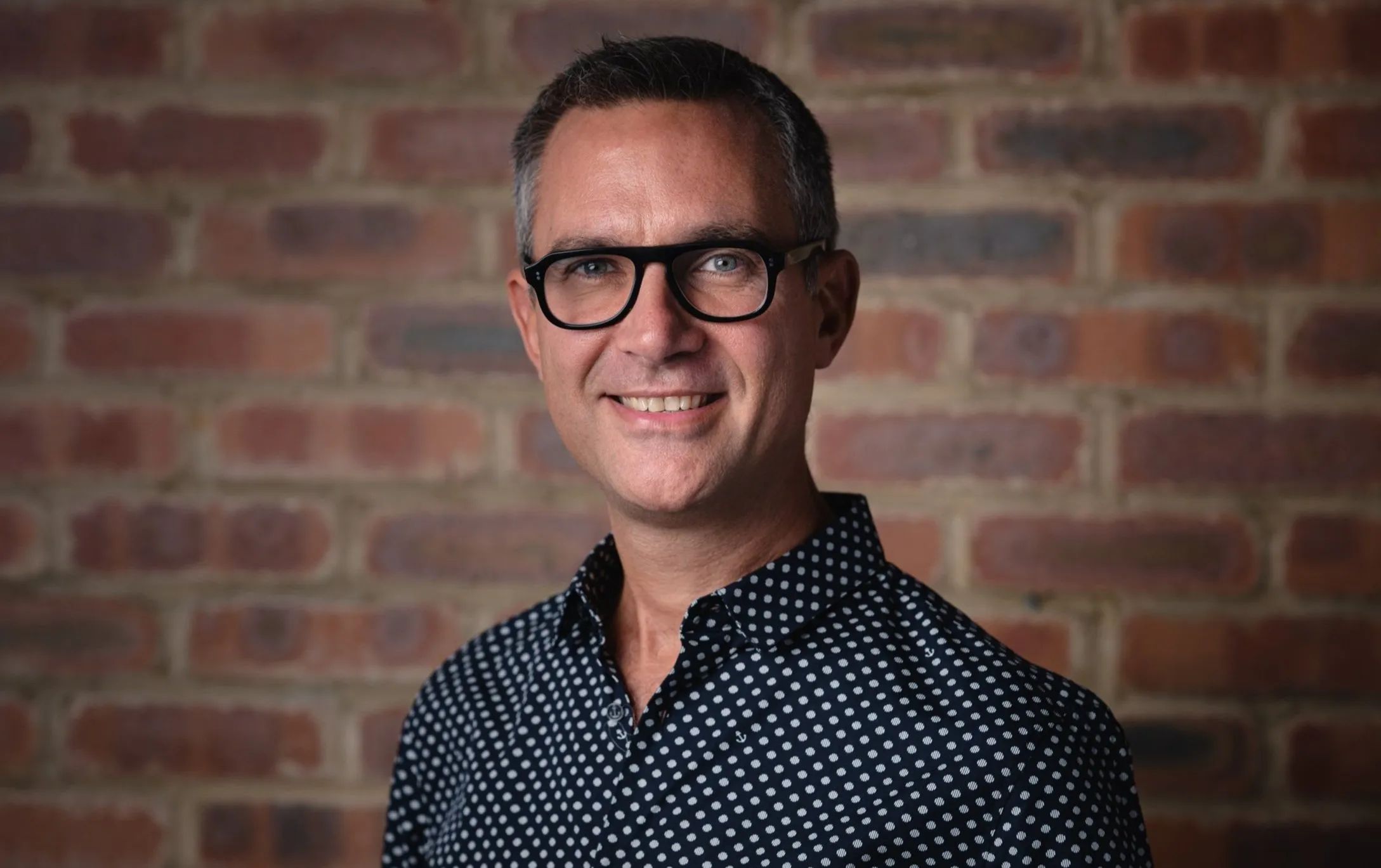 Creative corporate headshot of man in glasses and polka dot shirt, brick background, London
