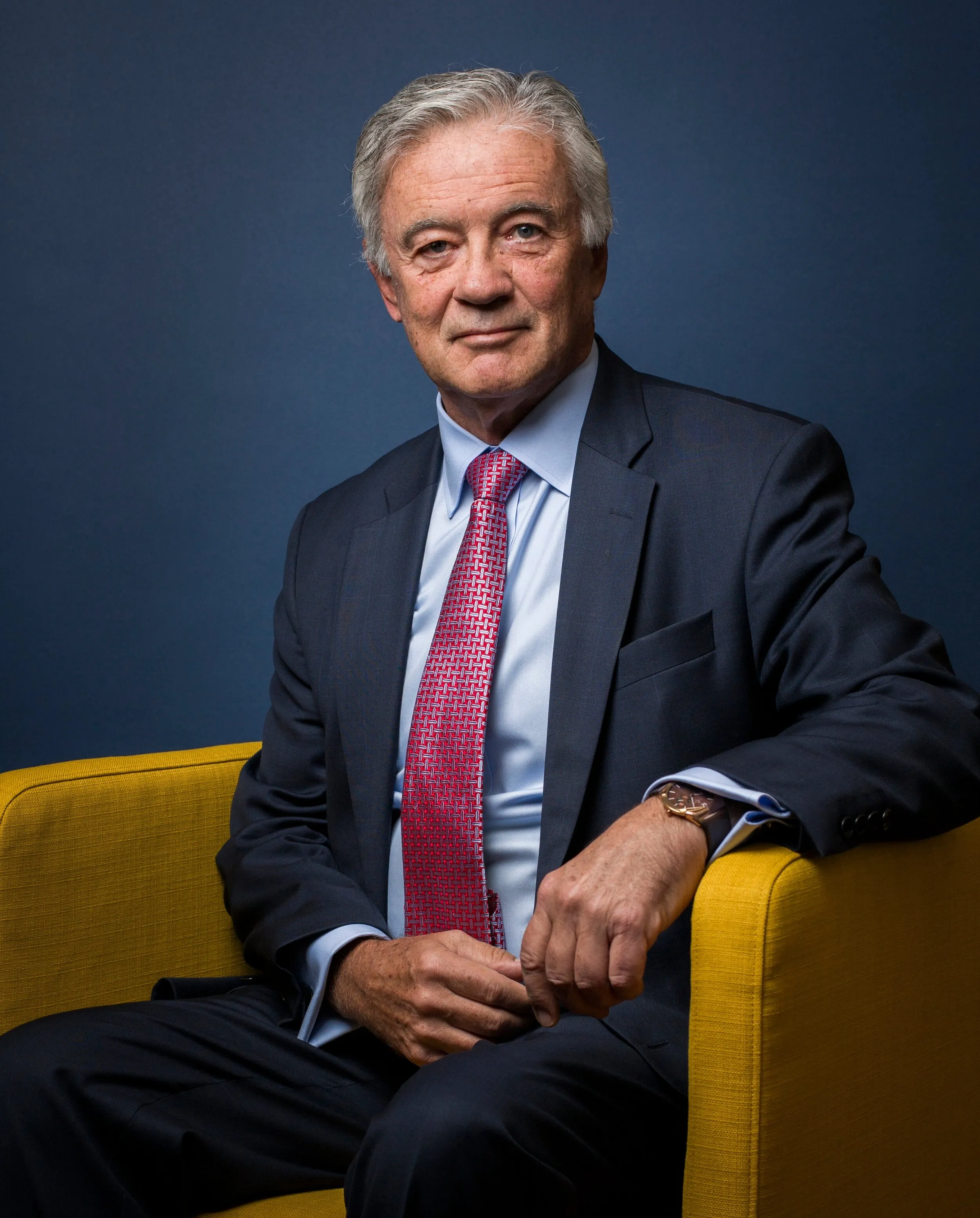 Senior executive headshot of man in navy suit with red tie, yellow chair, London