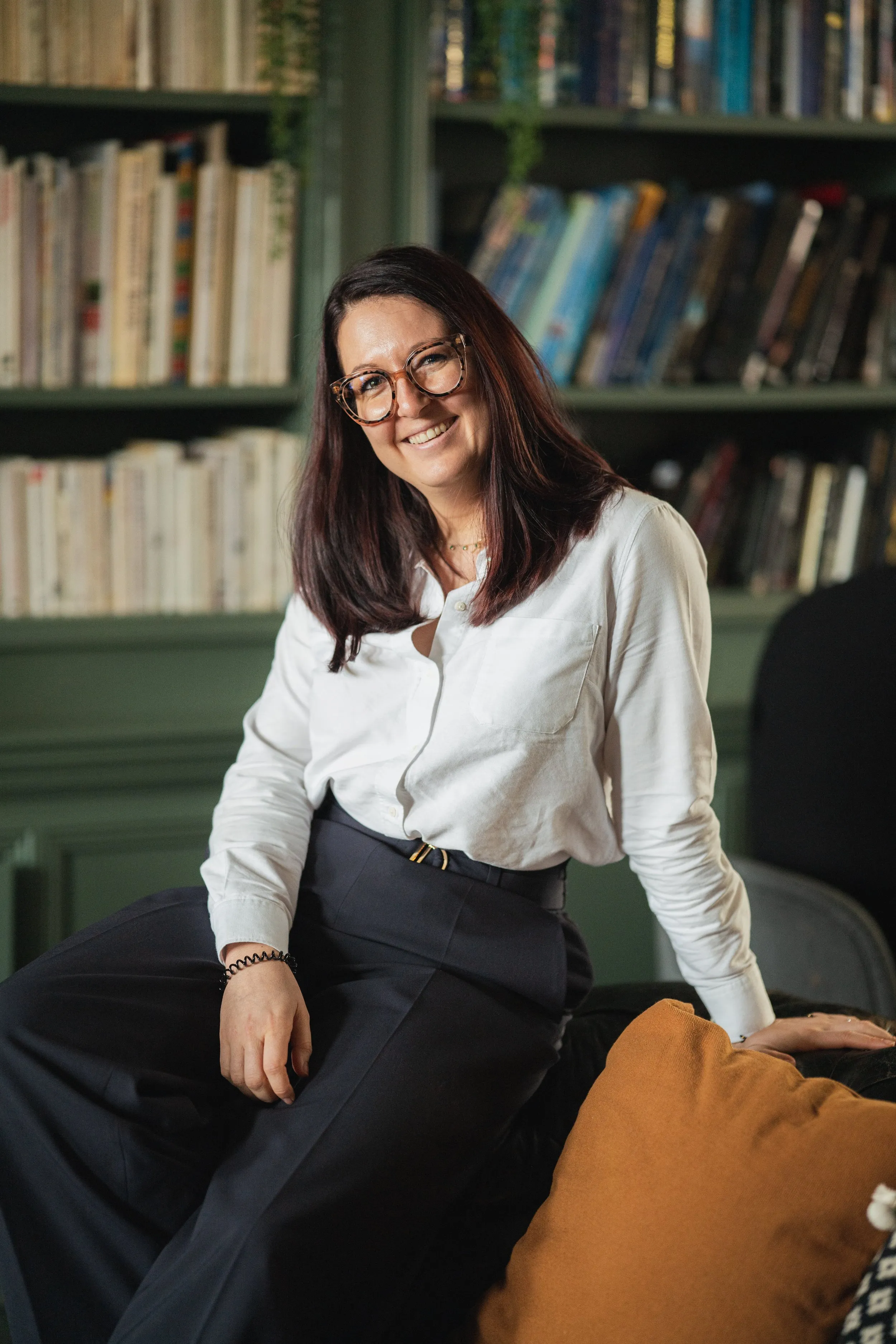 Professional headshot of woman in glasses and white shirt, bookshelf background, London