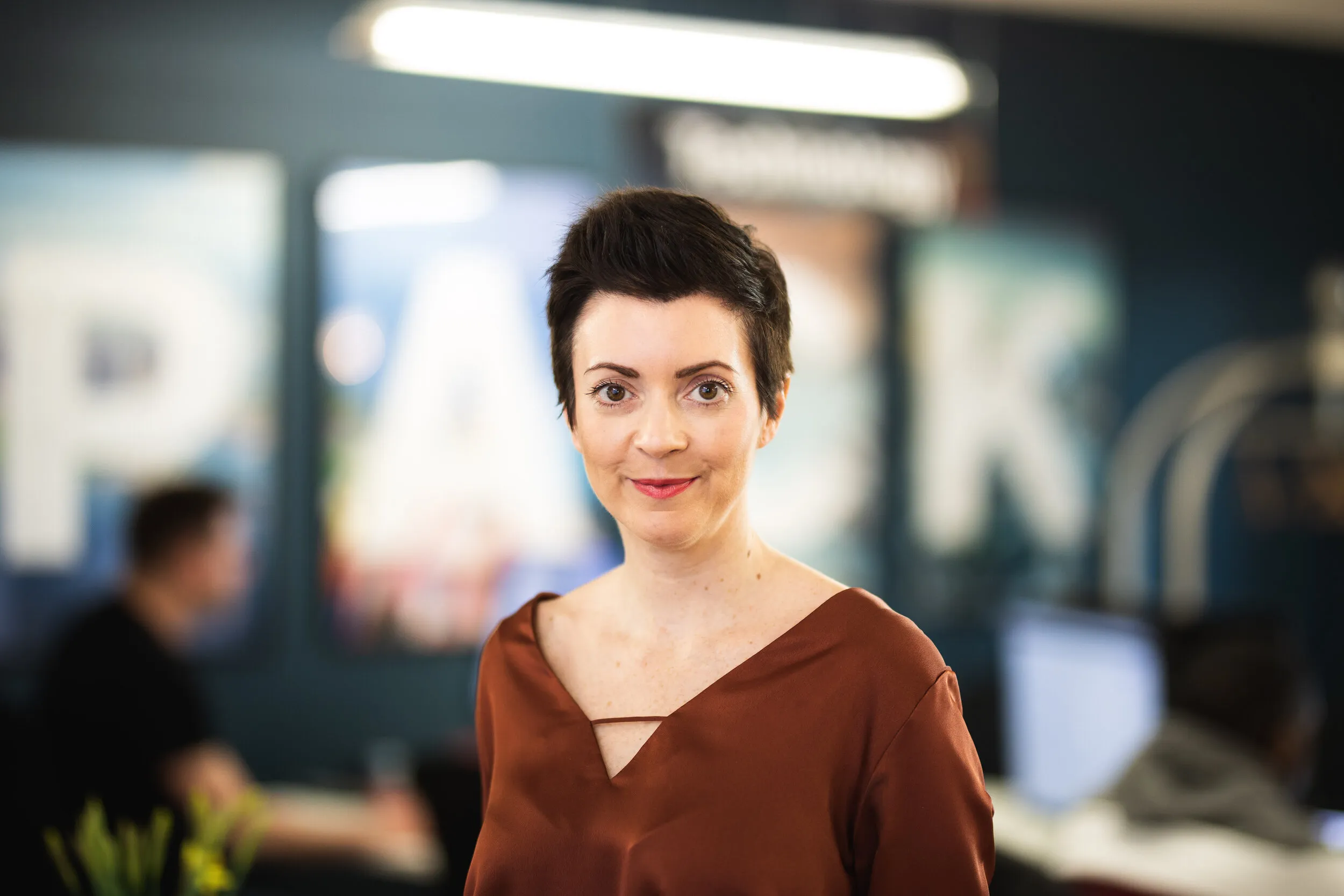 Professional headshot of woman with short dark hair in rust top, office background, London